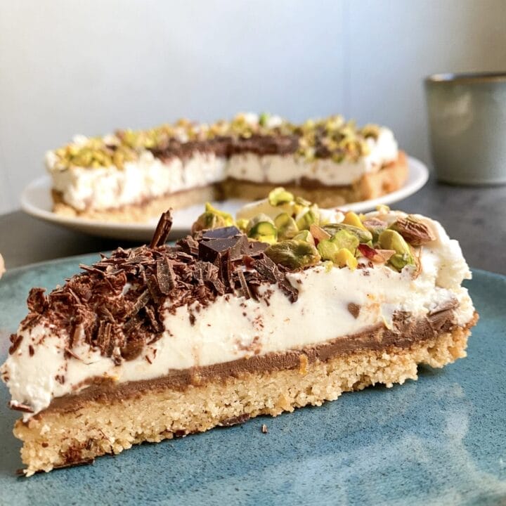 Picture of a slice of keto cannoli cheesecake on a blue plate. Cup of coffee and the rest of the keto cannoli tart in the background.